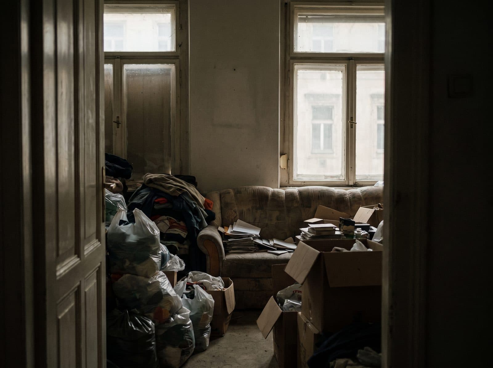 A neglected living room with piles of bags, clothes and boxes.